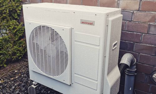 Weißes Außengerät einer Wärmepumpe der Marke Weishaupt vor einer Backsteinwand auf dunklem Schotter.