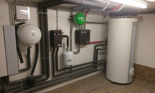 Technikraum mit Wärmespeicher, Verrohrung, Ausdehnungsgefäß und grüner Umwälzpumpe an gefliester Wand.