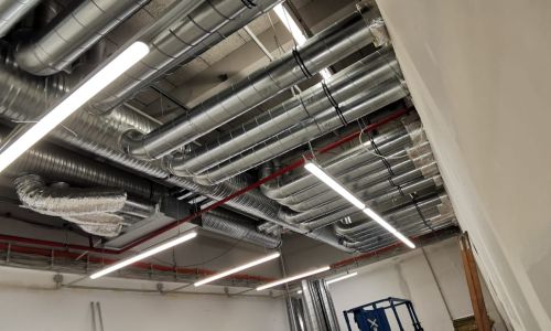 Industriedach mit zahlreichen Lüftungsrohren, Kabeltrassen und LED-Leuchten in einem Technikraum oder Rohbau.