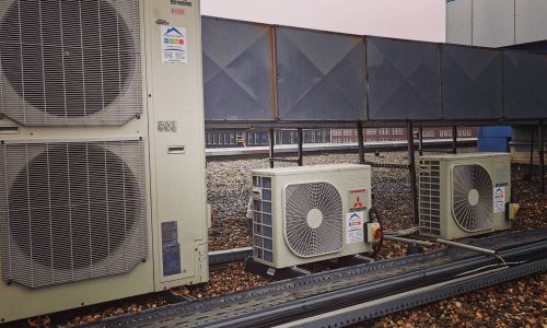 Drei Mitsubishi-Klimageräte auf einem Kiesdach, mit metallischer Umrandung und Gebäudefassade im Hintergrund.